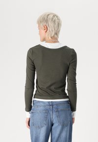 Pikkade varrukatega oliivroheline topp, millel on ümar kaelus, sooniktekstuur ja valge kihiline detail varrukaotsas. Kombineeritud helesiniste teksadega.