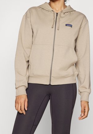 Persoon draagt een beige hoodie met rits en Patagonia-logo en donkere aangesloten legging, staand tegen een effen achtergrond.
