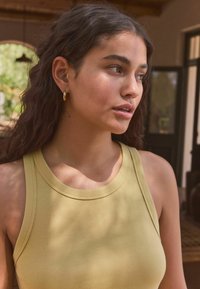Débardeur côtelé jaune clair avec un col rond et des bretelles larges. Le mannequin a de longs cheveux ondulés et porte de petites boucles d'oreilles en or.