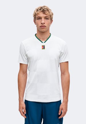 Jeune homme aux cheveux blonds courts et bouclés, portant un maillot de sport blanc à manches courtes avec un petit logo multicolore au col et un short bleu.