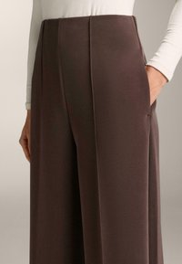 Hlače  širokih hlačnic iz temno rjave tkanine, z gladko teksturo, visokim pasom, čistimi linijami in stranskimi žepi. Vidni so subtilni detajli šivov.