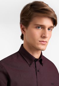 Giovane uomo con capelli castano chiaro che indossa una camicia botttonata di un profondo bordeaux scuro con un piccolo logo ricamato nero sul petto.