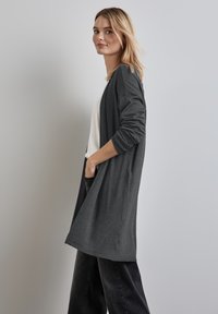 Blonde vrouw in een lange donkergrijze cardigan, wit topje en zwarte jeans staat zijwaarts met één hand in haar zak tegen een lichtgrijze achtergrond.