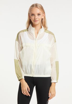 Jonge vrouw die een witte en lichtgroene windjack met rits en zwarte legging draagt, staand tegen een effen lichte achtergrond.