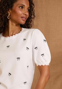 Baldai adīta blūze ar viļņotu kakla izgriezumu un puff piedurknēm, kurā visā virsmā ir melna loku izšuvuma. Tekstūras un viegla auduma.