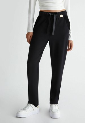 Woman wearing black drawstring pants with lace pocket details and white sneakers, standing against a plain light background.