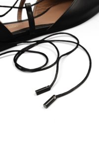 Black flat shoe with leather upper, featuring crisscross straps, slender ankle ties, and metallic tips on the shoelaces.