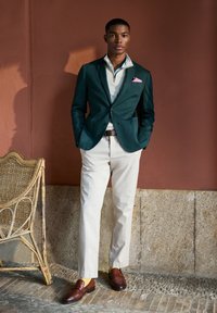 Blazer vert foncé sur une chemise de couleur claire, associé à un pantalon crème et des mocassins marron. La tenue comporte un mouchoir de poche rose.