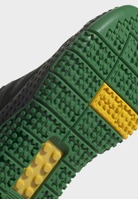 Sole černého tenisky s interlocking cihlovými vzory v zelené a žluté barvě, uspořádanými pro zajištění trakce.