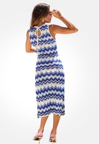 Mujer ajustándose las gafas de sol, vistiendo un vestido sin mangas de patrón ondulado en azul y blanco con un lazo en la espalda y sandalias de tacón beige.