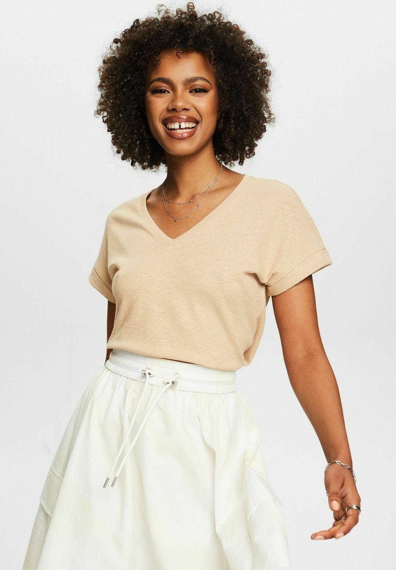Beige V-Ausschnitt-T-Shirt mit umgeschlagenen Kurzärmeln, kombiniert mit einem weißen Rock mit elastischem Bund und Zugband. Glatte Stofftextur.