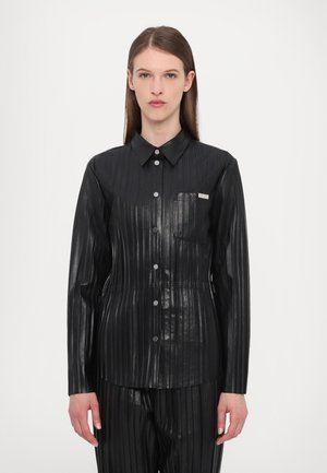 Camicia nera a pieghe in materiale lucido, dotata di colletto, chiusura frontale con bottoni e taschino sul petto. La texture appare liscia con linee verticali.