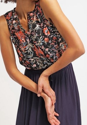 Kobieta w czarnej, bezrękawnikowej bluzce w czerwone i różowe kwiaty oraz ciemnej spódnicy z wysokim stanem, z rękami splecionymi na talii.