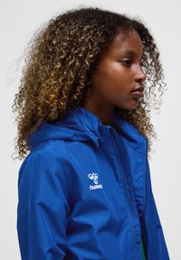 Jong persoon met krullend haar die een blauwe hummel-jas draagt, kijkt naar rechts tegen een effen lichte achtergrond.