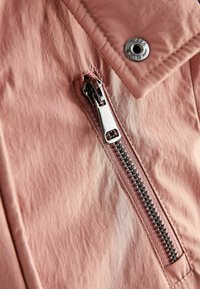 Veste rose clair en tissu texturé, avec une fermeture éclair argentée et un détail à bouton-pression sur le col. Ouverture zippée visible.