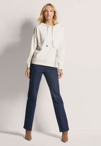 Crème hoodie met verstelbare trekkoorden, zijkant patroonaccenten, gecombineerd met donkerblauwe recht gesneden jeans en beige haklaarsjes.