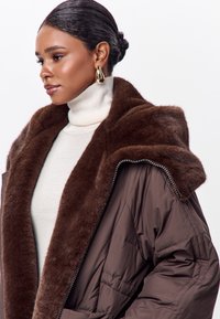 Chaqueta de puffer marrón con un gran y suave collar de piel sintética marrón, cierre con cremallera y tejido texturizado. Lleva puesta sobre un suéter de cuello alto color crema.