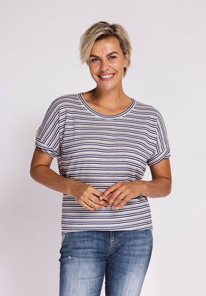 Lächelnde Frau mit kurzen blonden Haaren, die ein kurzärmeliges gestreiftes Hemd und blaue Jeans trägt, steht vor einem schlichten hellen Hintergrund.