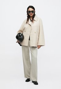 Μπεζ παλτό διπλό στερέωμα με φαρδιά μανίκια, μαύρη τσάντα, γκρι παντελόνι φαρδιάς γραμμής και μαύρα παπούτσια. Το μοντέλο φοράει σκούρα γυαλιά ηλίου.