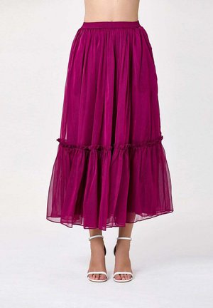 Longue jupe transparente fuchsia avec une couture froncée au-dessus de l'ourlet, portée avec des talons blancs à bride à la cheville, montrée de la taille aux pieds.