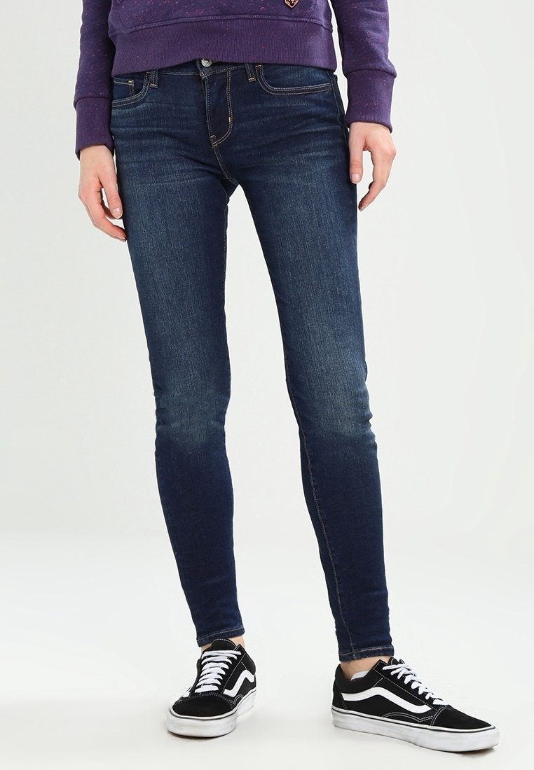 Pantalones vaqueros ajustados en denim azul oscuro con un ligero desvanecimiento. Combinados con zapatillas negro y blanco con rayas blancas. Un atuendo casual.