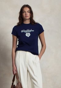 Tums zila kokvilnas T-kreklā ir balts "Wimbledon" logo un lauvas zīmējums priekšā. Īsās piedurknes un klasiskā piegriezte.