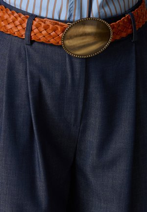 Gevlochten bruine leren riem met grote ovale messing gesp, gedragen over een donkerblauwe broek en een gestreept ingestopt overhemd.
