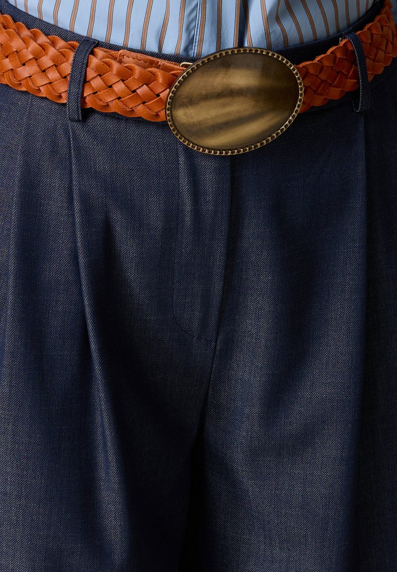 Cinturón trenzado de cuero marrón con hebilla grande ovalada de latón, llevado sobre pantalones azul oscuro y una camisa de rayas metida dentro.