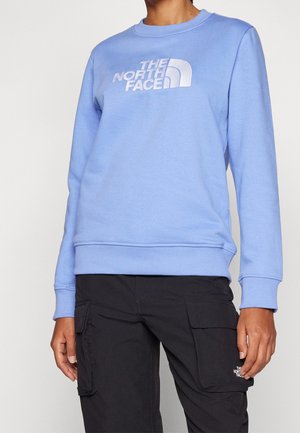 Person iført en lyseblå The North Face sweatshirt og sorte cargobukser med The North Face-logo på lommen.