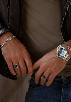 Silver-tone metal watch with black face, silver link bracelet, two silver rings with square accents, and a silver bar bracelet on a leather jacket background.