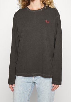 Frau trägt ein dunkelgraues Langarmshirt mit kleinem roten "N°21" Logo auf der Brust und hellblaue Jeans, steht vor weißem Hintergrund.