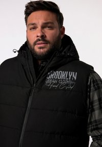 Svart vadderad väst med dragkedja fram och huva, med en texturerad grafisk tryck som visar "Brooklyn Urban Clothing" på bröstet.