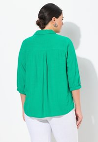 Grøn, let blouse med krave, trekvartærmer og plissert rygdesign. Kombineret med hvide bukser i en glat tekstur.