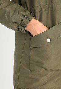 Olive green jacket with a textured finish, gathered cuff detail, and a side pocket featuring a snap button closure.