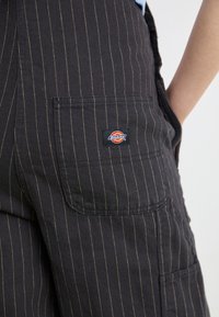 Schwarze Latzhosen mit goldenen Nadelstreifen, ausgestattet mit einer Seitentasche und einem schwarzen Dickies-Logo-Patch. Texturiertes Baumwollmaterial.