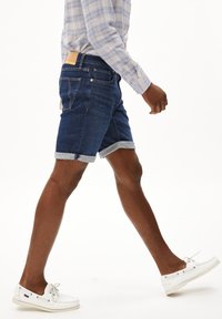 Donkerblauwe denim shorts met opgerolde zomen, voorzien van een traditionele vier-pocketontwerp en een lichtbruine leren label op de tailleband. Draagt deze met witte schoenen.