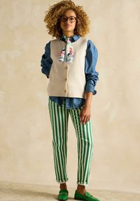 Beige Weste aus Wolle mit Knöpfen über einem Jeanshemd, kombiniert mit grün-weiß gestreiften Hosen und grünen Loafers. Der Schal hat ein florales Muster.
