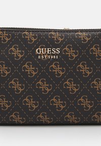 Portefeuille en cuir marron foncé avec motif répétitif du logo Guess et inscription métallique Guess EST 1981 sous la fermeture éclair.