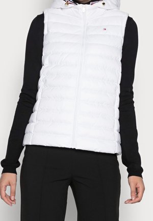 Osoba ubrana w białą pikowaną kamizelkę bez rękawów z zamkiem z przodu i kapturem, czarną bluzkę z długim rękawem oraz czarne spodnie.