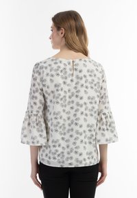 Blomstret bluse i cremefarve med sorte mønstre, trekvart bellærmer, nøglehul detalje bagpå og let stof. Sorte bukser på.