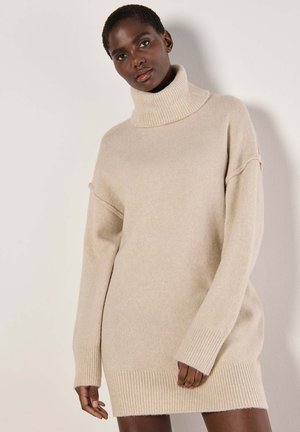 Robe en tricot beige avec un col roulé haut, coupe oversize et poignets et ourlet côtelés, présentant une texture douce.