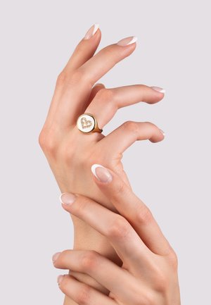 L'anello d'oro presenta una faccia circolare con un design a cuore in rilievo decorato con gemme. Le mani hanno una finitura naturale e una leggera manicure francese.
