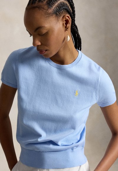 Γυναίκα με πλεξουδάκια, φορώντας ανοιχτό μπλε πλεκτό τοπ με κοντά μανίκια και μικρό κίτρινο κεντητό λογότυπο, καθώς και σκουλαρίκι με μαργαριταρένιο σταγόνα, κοιτάζει προς τα κάτω.