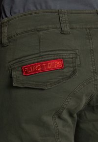 Pantalon vert olive avec un rabat de poche arrière présentant un patch brodé rouge portant l'inscription "FLYING TIGERS".