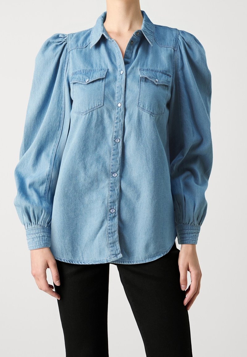 Chemise en denim bleu clair avec manches longues bouffantes, col à revers, fermeture à boutons sur le devant et deux poches poitrine. Texture lisse.