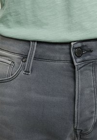 Vaqueros de mezclilla gris con una textura suave, que presentan un cierre frontal de botón, trabillas para el cinturón y bolsillos a los lados. Camisa visible arriba.