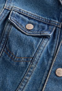 Tissu en denim bleu avec des surpiqûres orange détaillées et des boutons métalliques portant l'inscription « Authentic 1982 Next Dude » sur un rabat de poche.