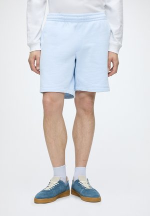 Mann trägt hellblaue Shorts, weißes Langarmshirt, weiße Socken und blaue Turnschuhe mit Gummisohlen, steht vor einem schlichten Hintergrund.