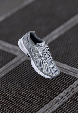 Chaussure de sport New Balance gris et blanc avec une tige en mesh reposant sur une surface en grille métallique.
