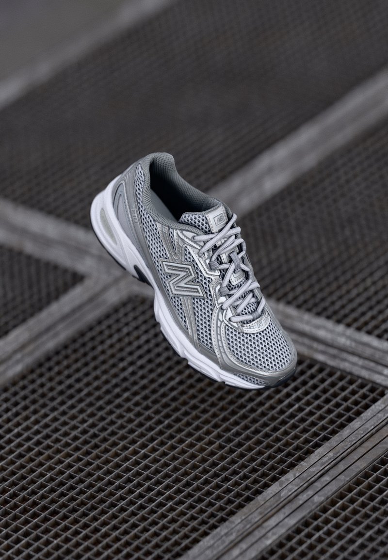 Chaussure de sport New Balance gris et blanc avec une tige en mesh reposant sur une surface en grille métallique.